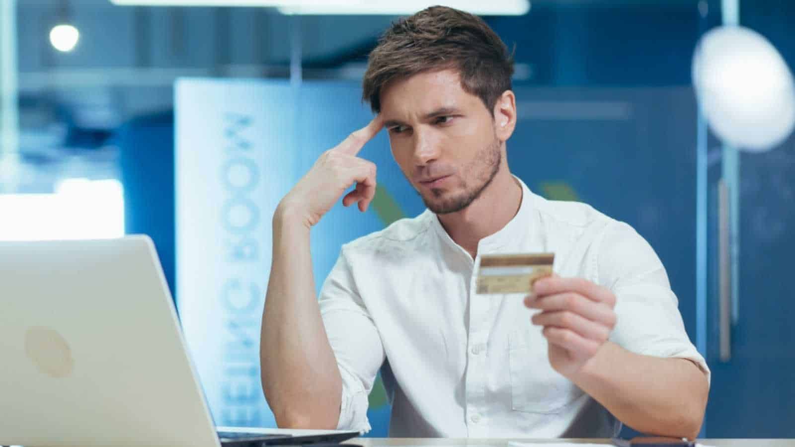 Pensive and frustrated young businessman in the office trying to make a purchase in an online store, uses a laptop holding a bank credit card