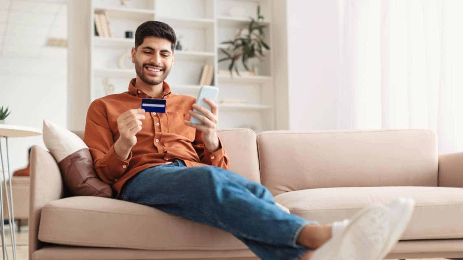 Internet Mobile Shopping Concept. Positive Middle Eastern male using cell phone holding looking at credit card purchasing things online sitting on sofa buying gifts in web store, full body length